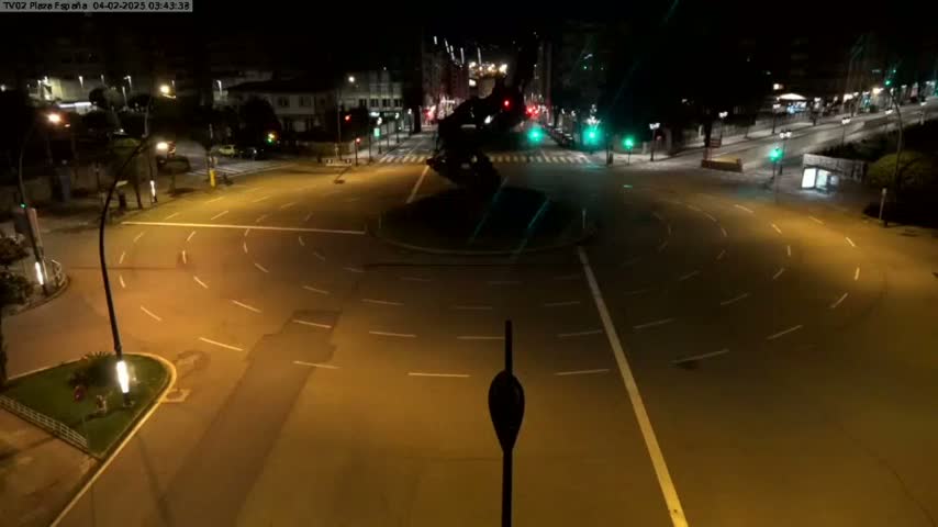 An empty roundabout at night, illuminated by streetlights, is surrounded by quiet roads and buildings.