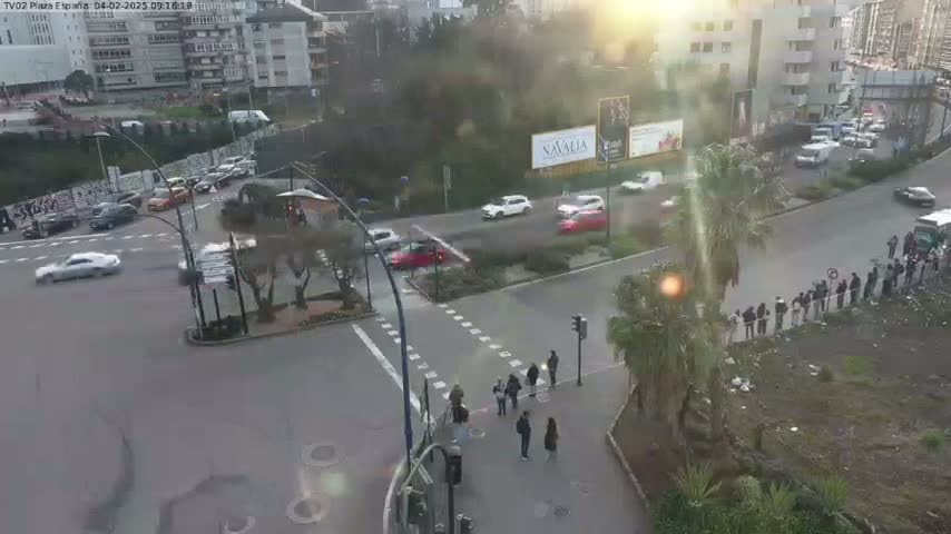 A sunny day shows a bird's-eye view of a city intersection with several cars at a crosswalk and a group of people standing along a curb.