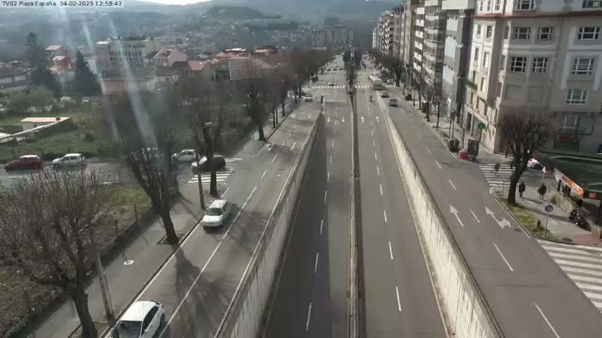 A sunny day view of a multi-lane road, partially divided by a high concrete wall, with cars driving in both directions, and buildings and trees lining the sides of the road.