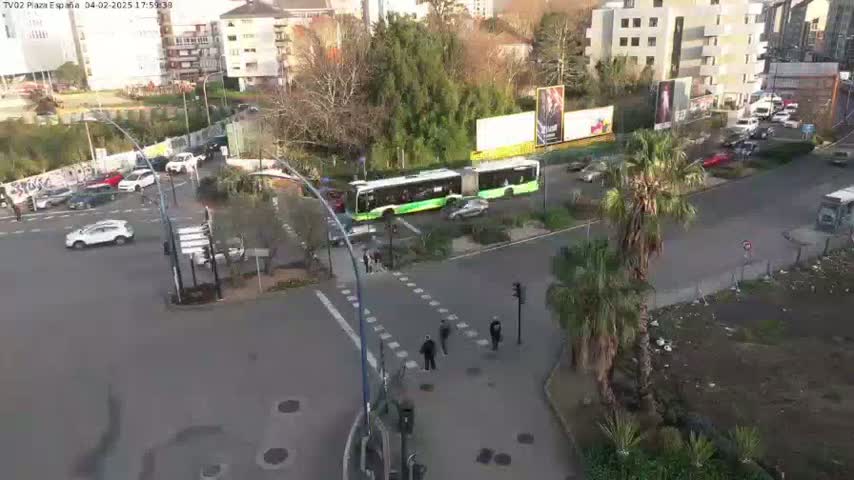 A high-angle view shows a city intersection with several cars and a long green bus,  under a mostly clear sky.