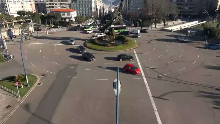 A sunny day shows several cars navigating a roundabout in a city center,  surrounding a central island with a statue and landscaping.