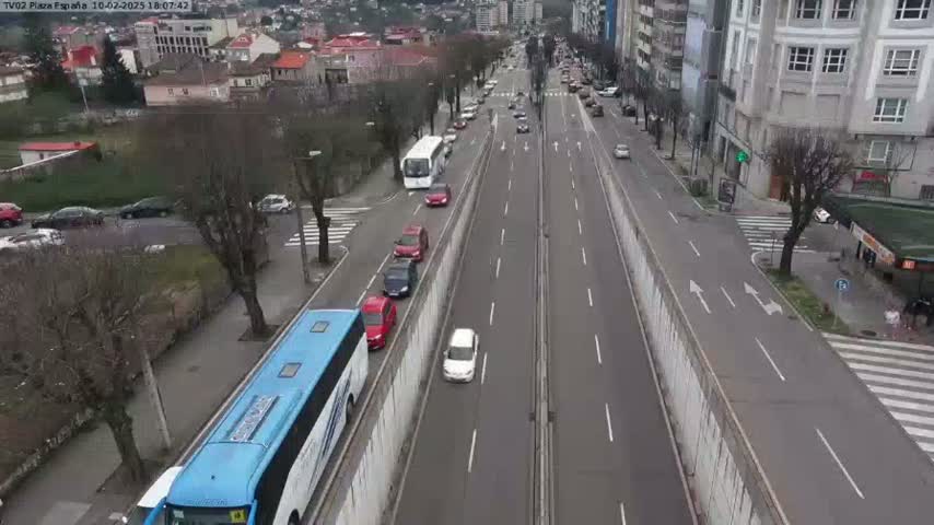A long, divided highway with several cars and buses driving in both directions, bordered by buildings and trees on an overcast day.
