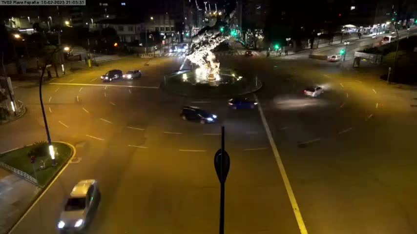A nighttime aerial view shows several cars navigating a roundabout centered around a fountain-like structure, illuminated by streetlights.