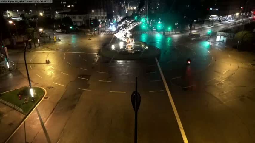 A wet, nighttime view of a roundabout in a city, illuminated by streetlights, with few vehicles present.