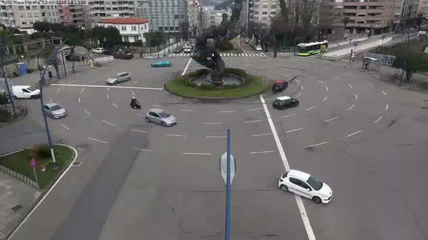 A roundabout with several cars and motorcycles navigating it under a cloudy sky.