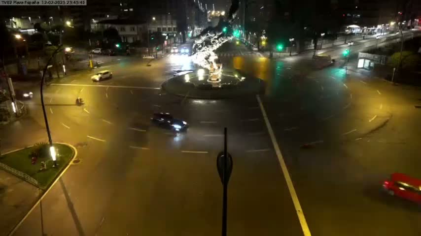 A nighttime, wet road intersection with a fountain in the center, showing several cars traveling around the roundabout.