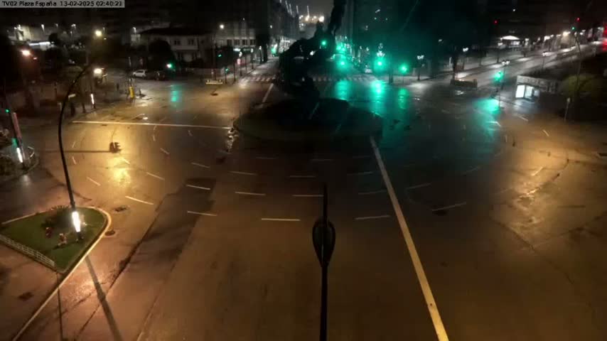 A mostly empty, wet roadway intersection at night, illuminated by streetlights and traffic signals.