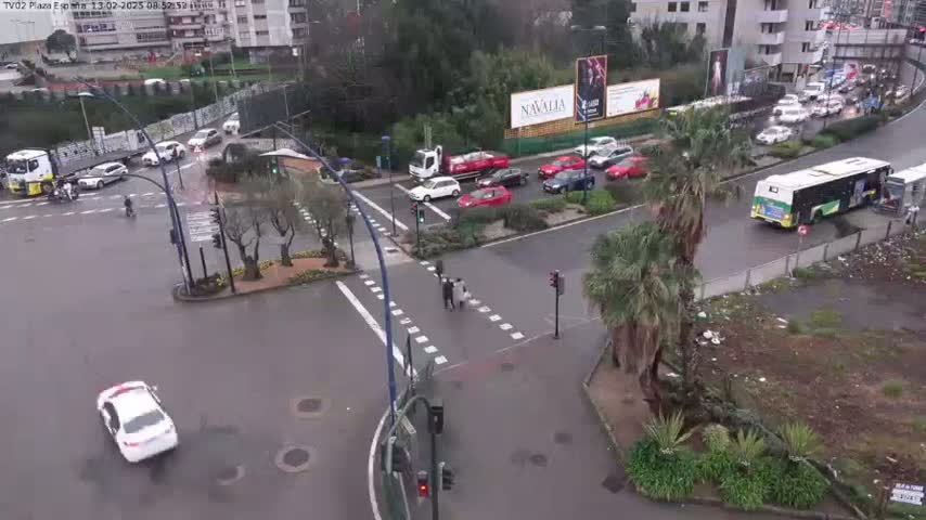 A wet, urban intersection shows several cars and a bus in motion, with pedestrians crossing a crosswalk.