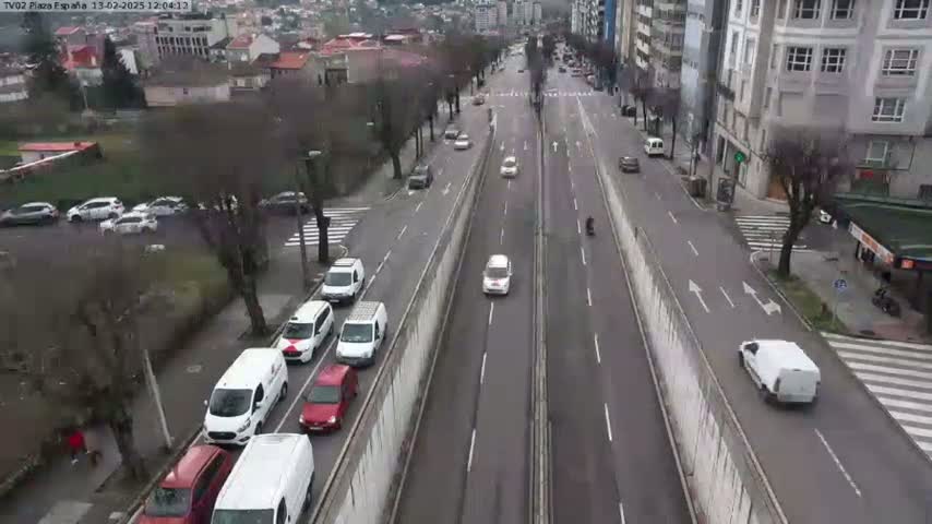 A highway scene shows numerous cars and vans driving in both directions on a multi-lane road, partially divided by a concrete barrier, on an overcast day.