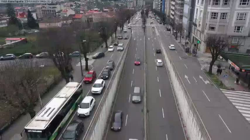 A long, multi-lane road with moderate traffic flows in both directions, bordered by buildings and trees on either side, under a cloudy sky.