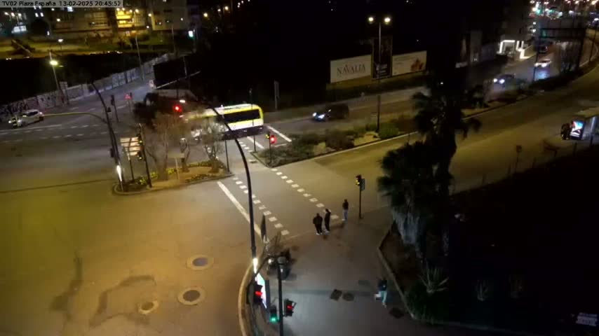 A nighttime street scene shows a yellow bus and several cars at an intersection, with a few pedestrians near a crosswalk and palm trees lining the road.