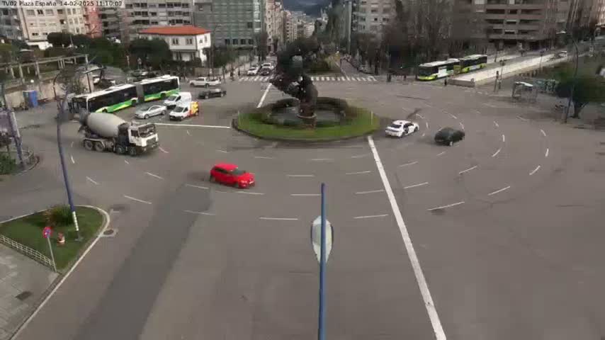 A cement mixer truck and several cars navigate a roundabout in front of a large sculpture, on a mostly clear day.