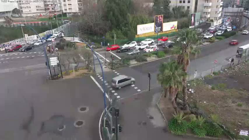 A wet, city intersection with several cars stopped at traffic signals and other vehicles moving along a multi-lane road, surrounded by buildings and greenery.