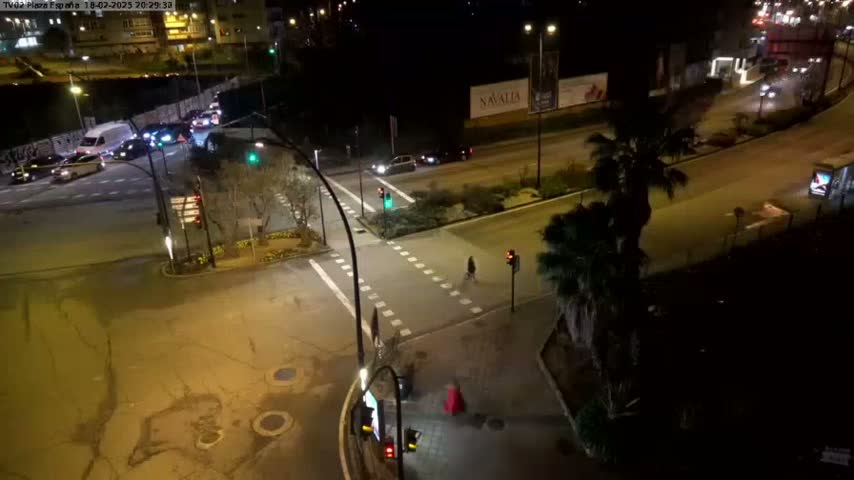 A nighttime aerial view of a multi-lane road intersection with sparse traffic and pedestrians, illuminated by streetlights.