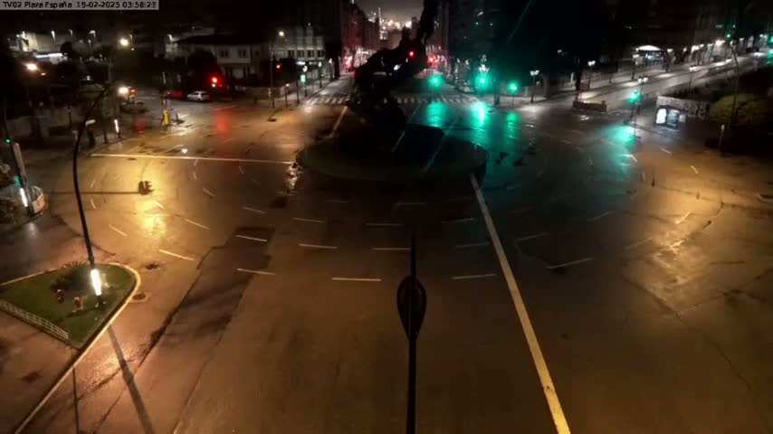 A wet, empty roundabout and intersecting roads are illuminated at night by streetlights.