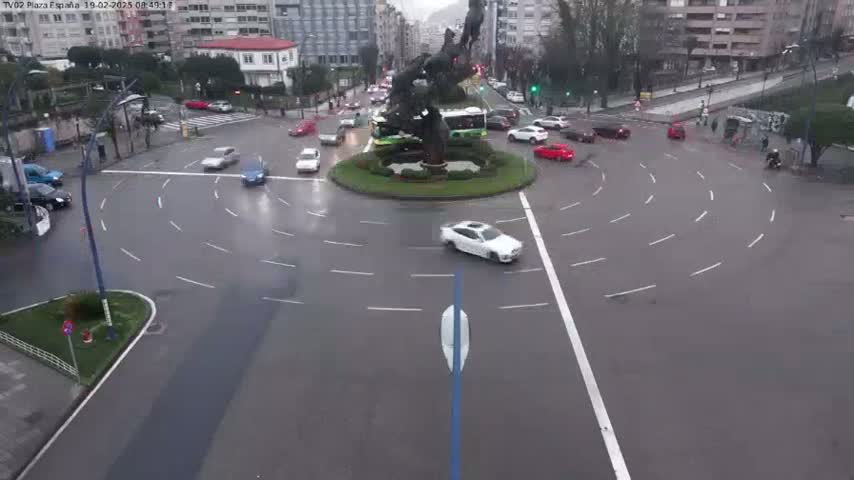 A wet roundabout in a city, with several cars navigating its lanes and a large statue in its center.