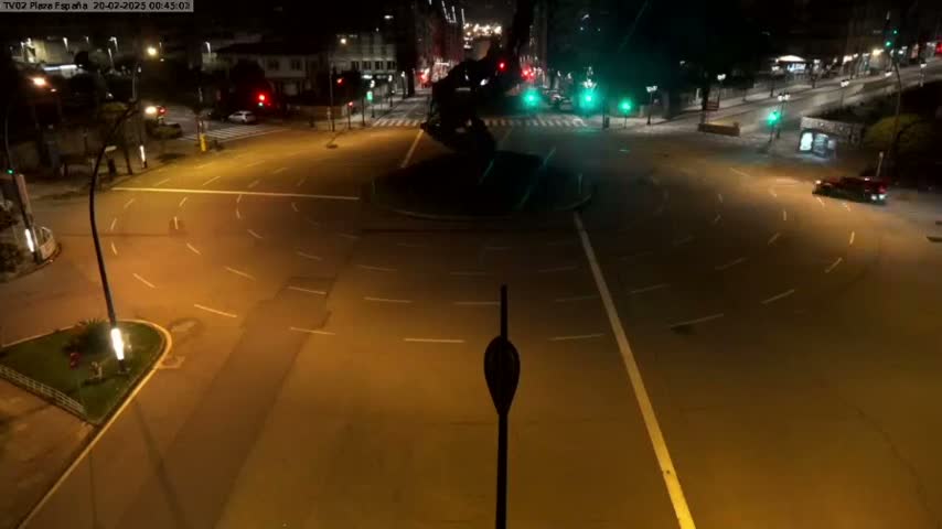 A mostly empty roundabout at night, illuminated by streetlights, with a few cars visible in the distance.