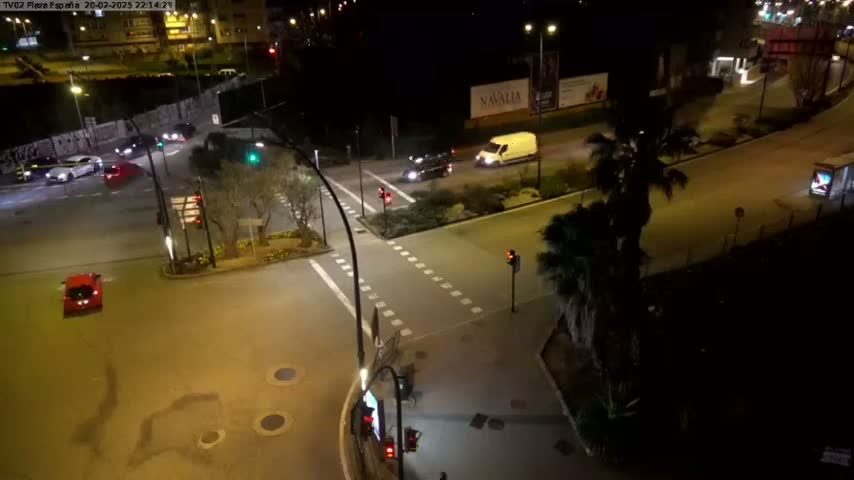 A nighttime aerial view shows several cars driving through a multi-lane intersection with traffic signals, surrounded by palm trees and buildings.