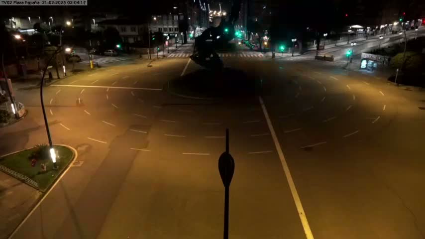A mostly empty, circular roadway at night is illuminated by streetlights, featuring a dark, statue-like structure in its center.