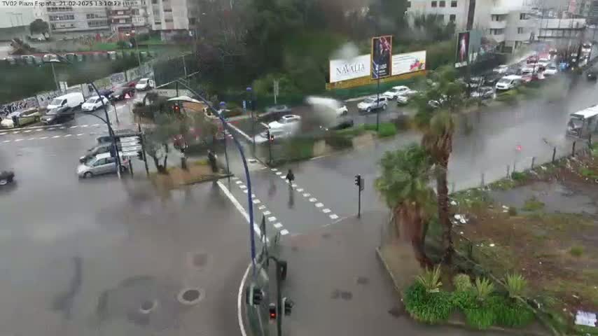 A rain-soaked street intersection shows several cars navigating the wet roads, pedestrians crossing, and palm trees swaying in the downpour.