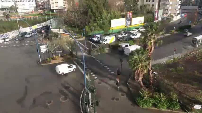 A wet, city street intersection is viewed from above, showing several vehicles, pedestrians, and palm trees, with buildings and a billboard in the background.