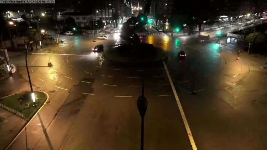 A rain-slicked roundabout at night, with several cars navigating its circular path and surrounding streets under streetlights.