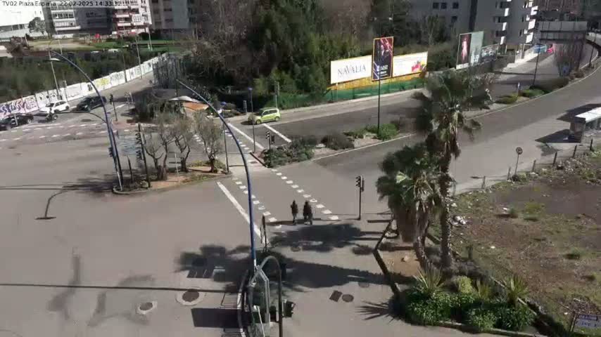 A sunny day shows a street intersection with some pedestrians, a few cars, palm trees, and buildings in the background.