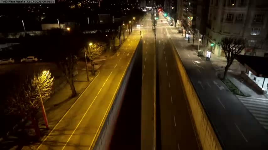A mostly empty, brightly lit roadway at night is divided by a high wall, with sparsely populated sidewalks and buildings lining either side.