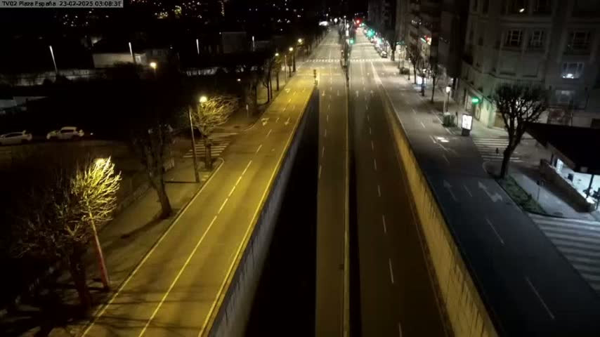 A mostly empty, well-lit city street at night, divided by a dark central barrier, with a few parked cars visible in the distance.