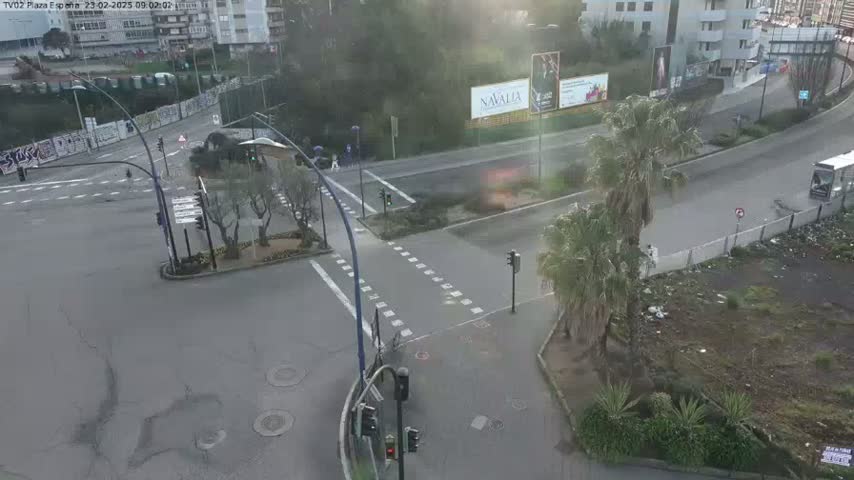 An aerial view of a street intersection with traffic signals, palm trees, and buildings in the background on a slightly overcast day.