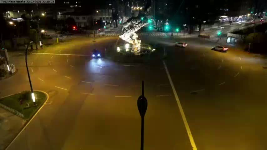 A nighttime, overhead view of a roundabout with several cars and a central fountain.
