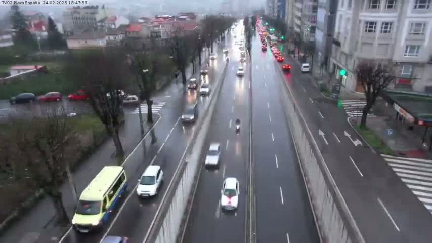 A wet, multi-lane road with moderate traffic is viewed from an elevated position, showing cars and a van traveling in both directions, with buildings and trees visible on either side.