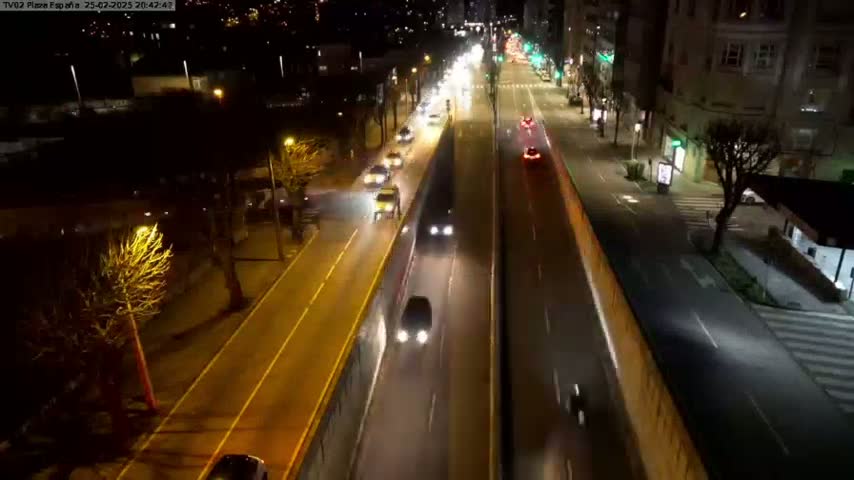 A nighttime, high-angle view shows a multi-lane road with cars driving in both directions, lined with trees and buildings.