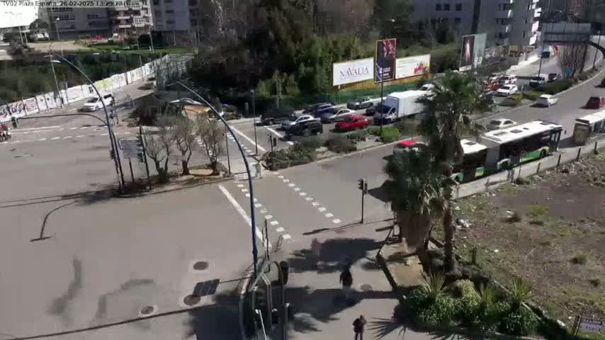 A sunny day shows a street intersection with several cars and buses, pedestrians, and some buildings in the background.