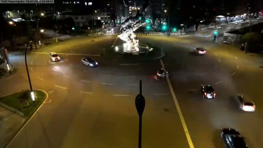 A nighttime aerial view shows several cars navigating a roundabout centered around a water feature, illuminated by streetlights.