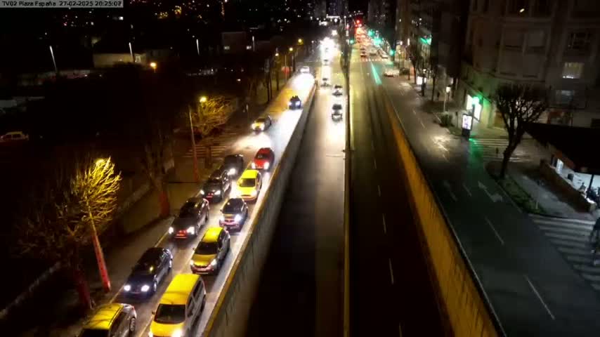 A nighttime, high-angle view of a city street with a line of cars driving slowly in one lane, alongside a higher-elevation roadway with sparse traffic.