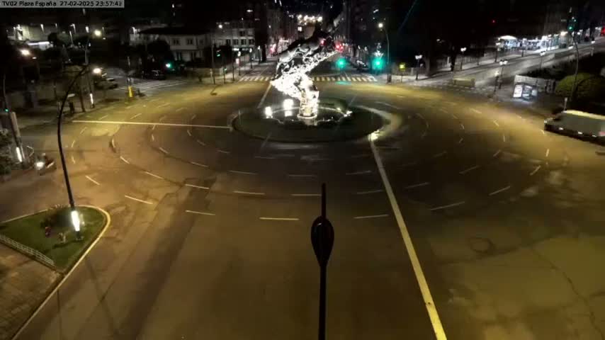 A nighttime aerial view shows a circular traffic roundabout with a fountain in the center, surrounded by city streets and buildings under clear skies.