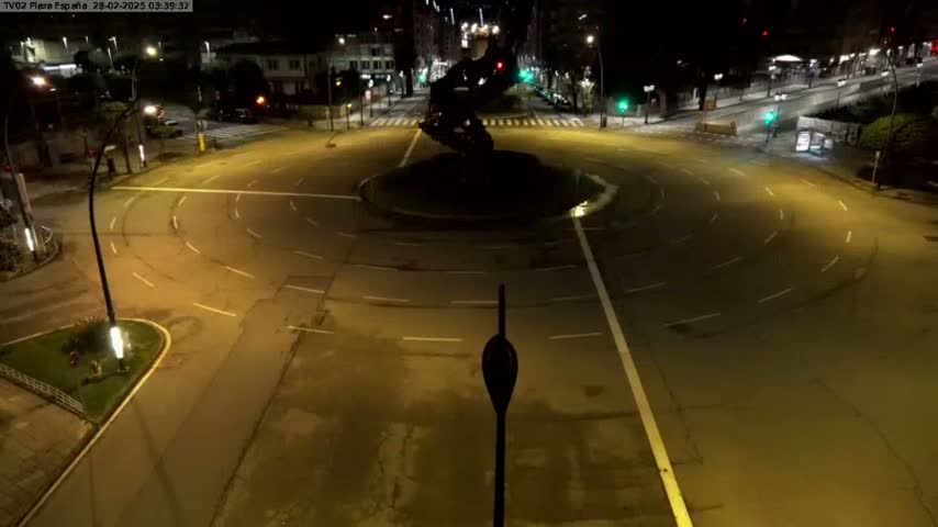 An empty roundabout at night, illuminated by streetlights, is surrounded by buildings and roads.