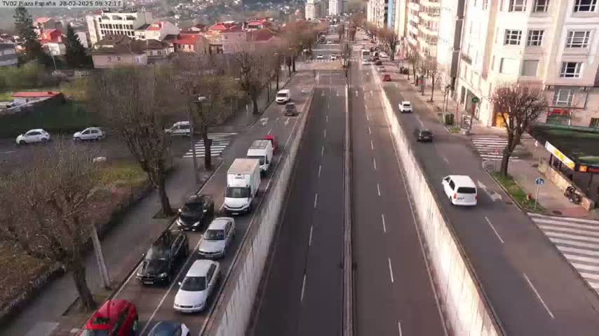 A high-angle view of a city street with parked and moving cars on a sunny day, alongside buildings and trees.