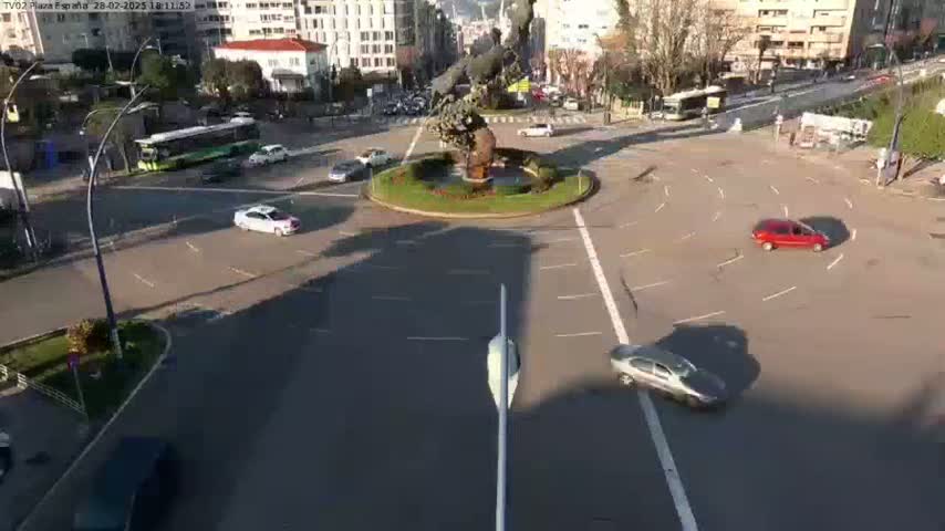 A sunny day shows several cars driving around a traffic circle featuring a large statue, with buildings and buses visible in the background.