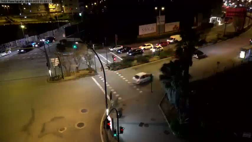 A nighttime aerial view of a city intersection with several cars at a traffic light and several more cars driving on the roadways.