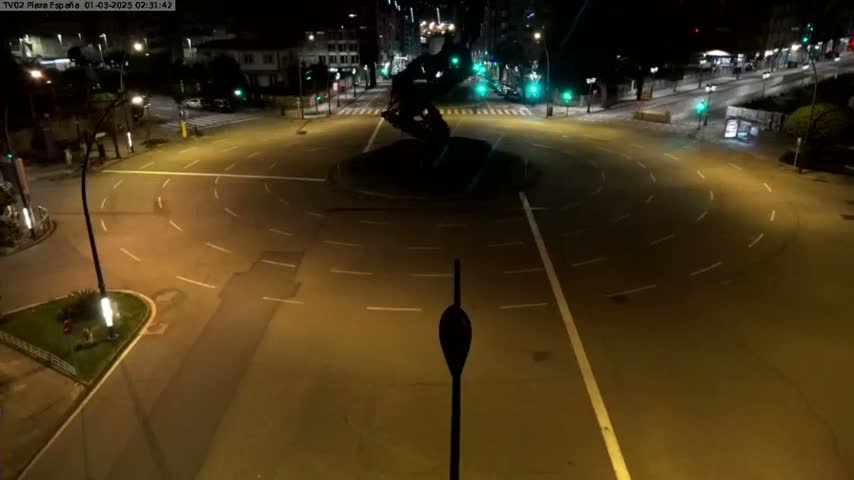 A mostly empty roundabout at night, illuminated by streetlights, with a dark sculpture in the center.