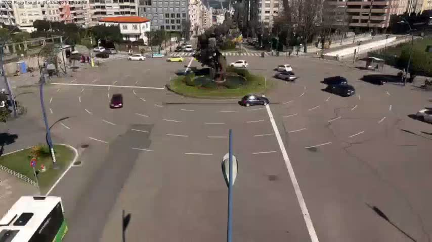 A roundabout with several cars driving around a central fountain on a sunny day.