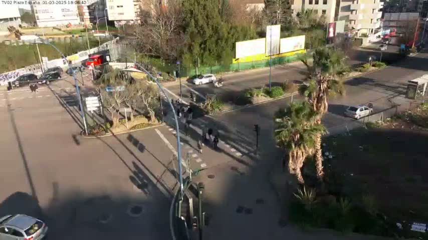 An aerial view of a sunny city intersection shows several cars and pedestrians, with palm trees and other landscaping visible.
