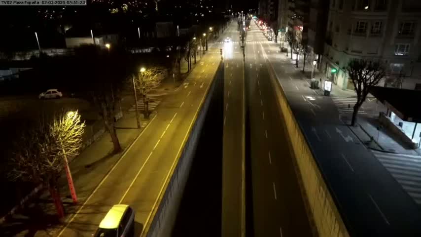 A nighttime aerial view of a mostly empty, brightly lit city street divided by a wall, with a few cars visible.