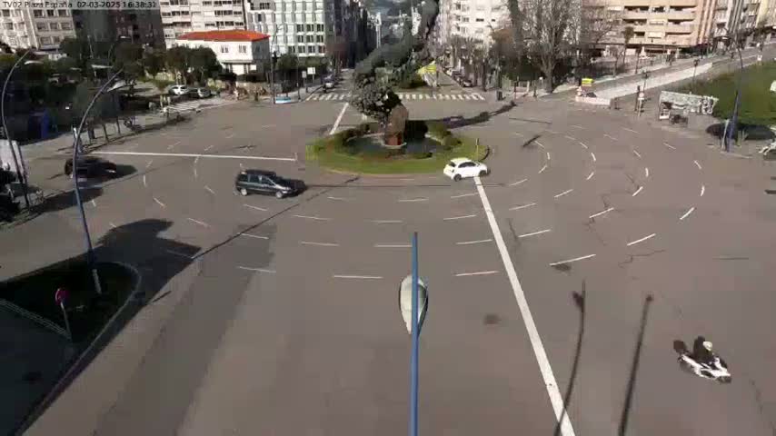 A sunny day shows a roundabout with several cars and a person on a scooter in a city center.