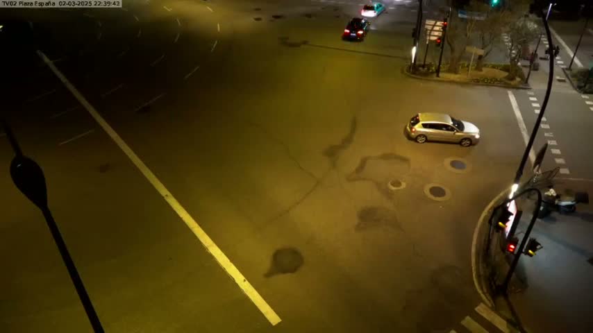 A nighttime, high-angle view shows a street intersection with a few cars, including a silver car driving through the intersection.