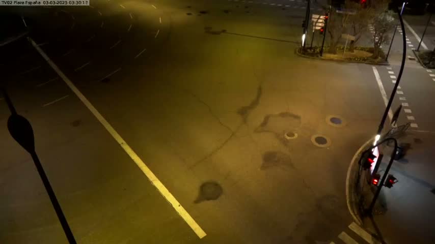 An empty, dark intersection at night shows several manhole covers and streetlights illuminating wet pavement.