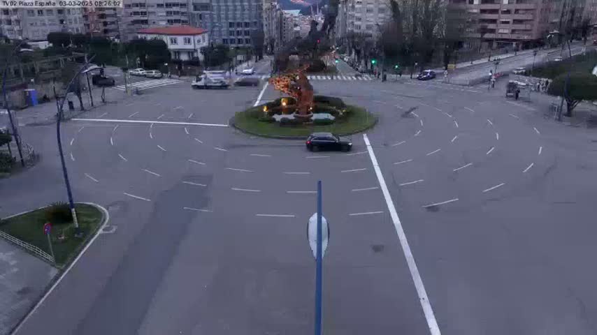 A mostly empty roundabout in a city, with a few cars and buildings visible in the background, on an overcast day.