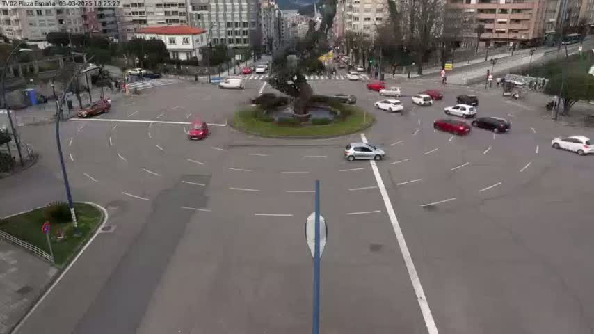 A traffic circle with several cars circulating around a central monument, under a partly cloudy sky.